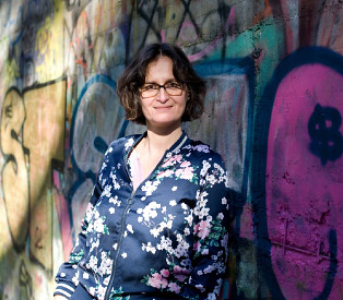 Rabea Wienholt mit bunter Jacke, lehnt an einer Graffiti-Wand